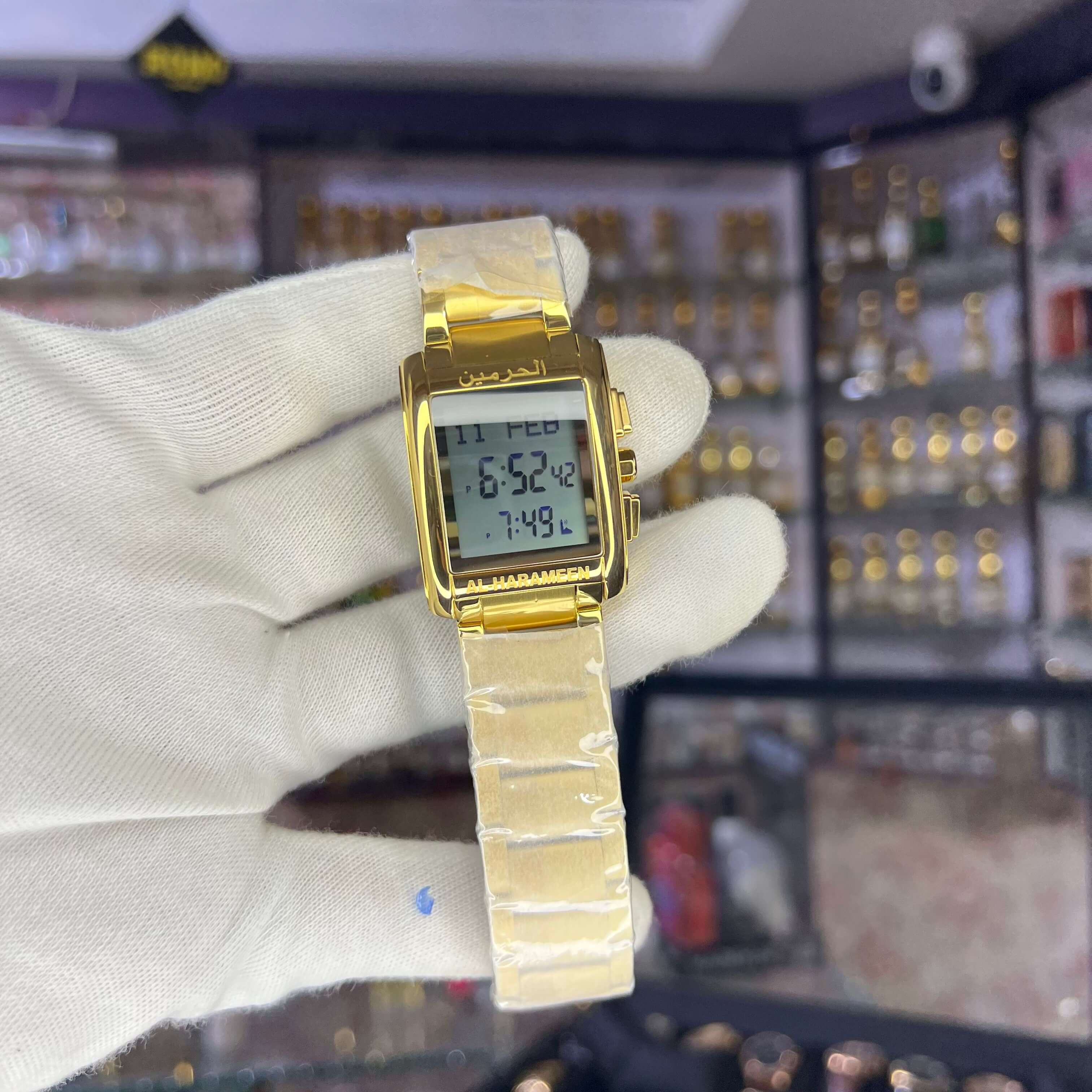 Gold digital watch held by a gloved hand with a blurred background of jewelry store.