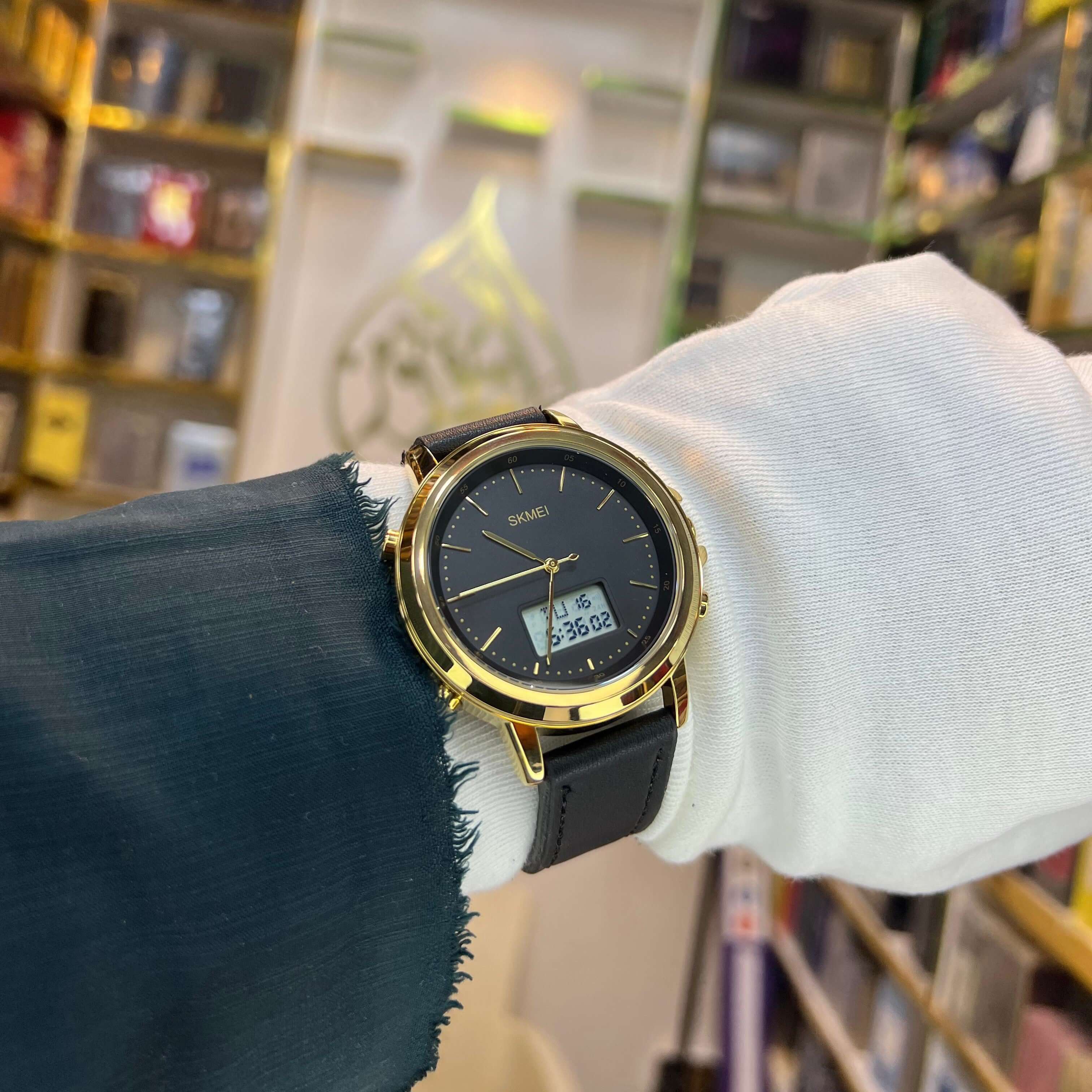 Gold wristwatch with black strap on a person's wrist, blurred bookshelf background