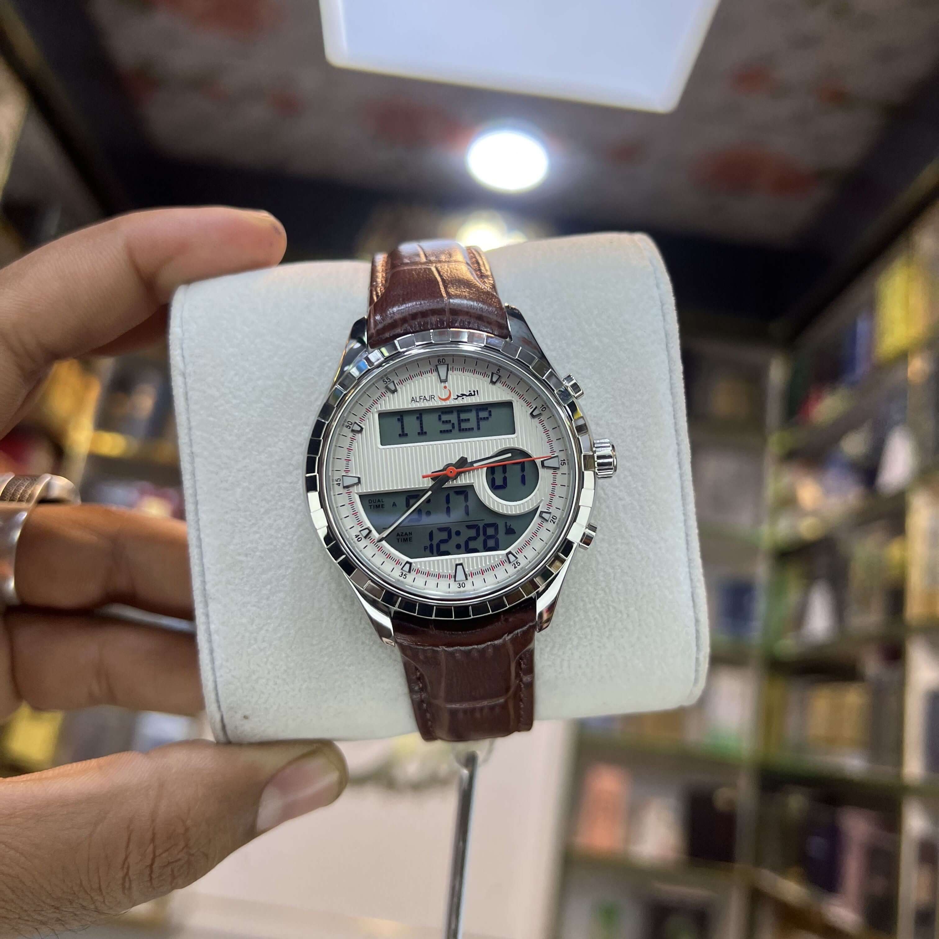 Digital wristwatch with brown leather strap on a white display stand, held by a hand.