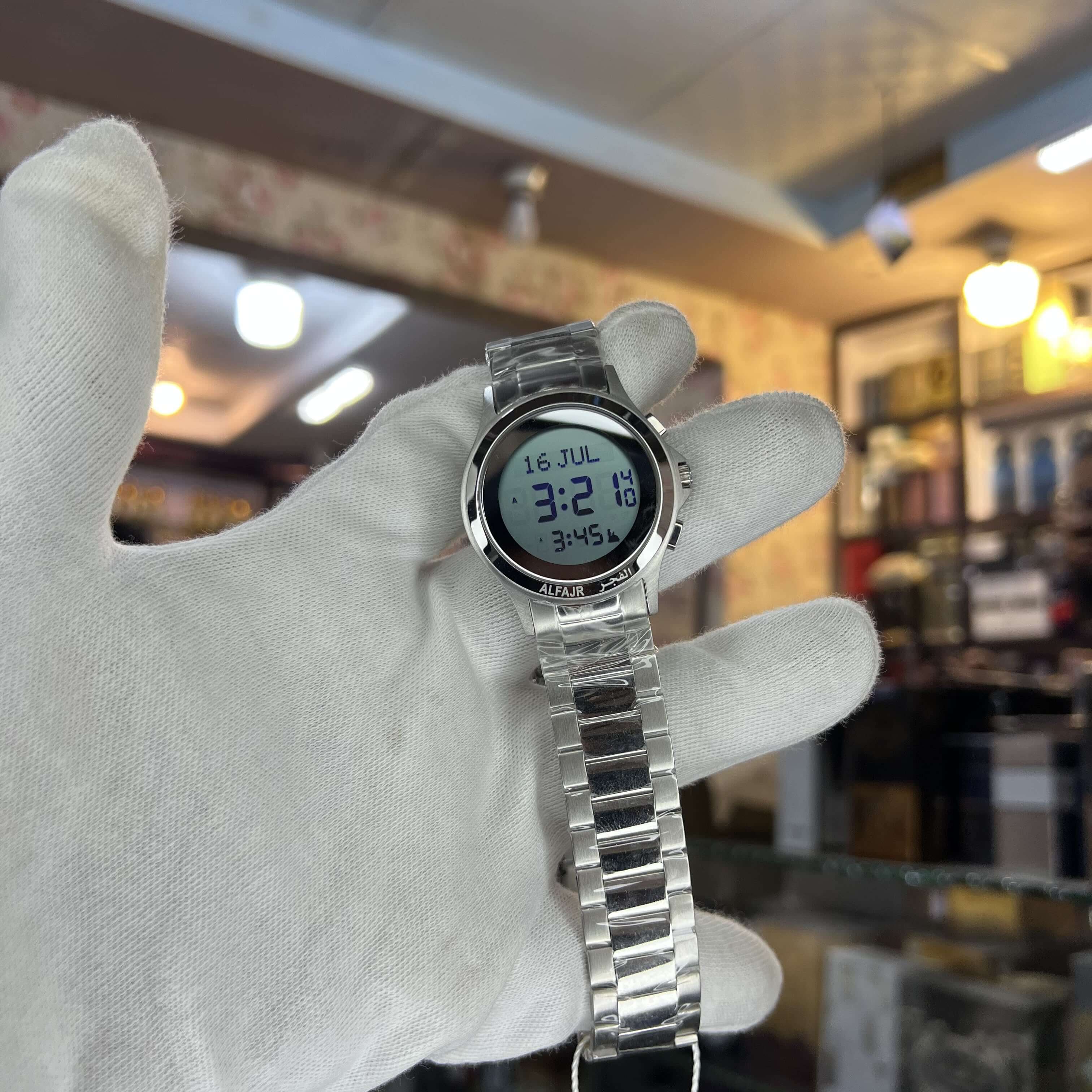 Silver digital watch held in a white gloved hand with a blurred indoor background