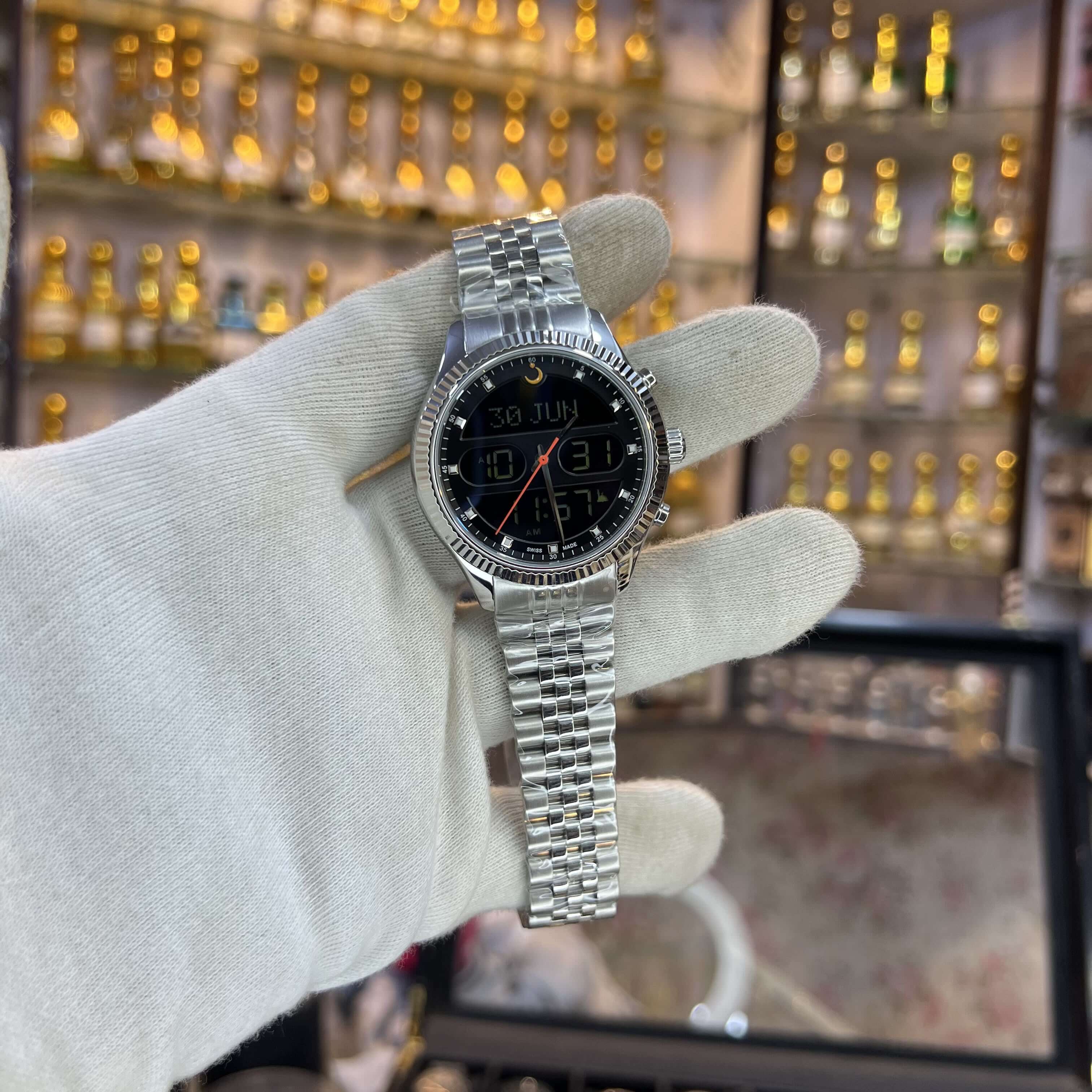 Silver watch held by a gloved hand with a display of perfume bottles in the background