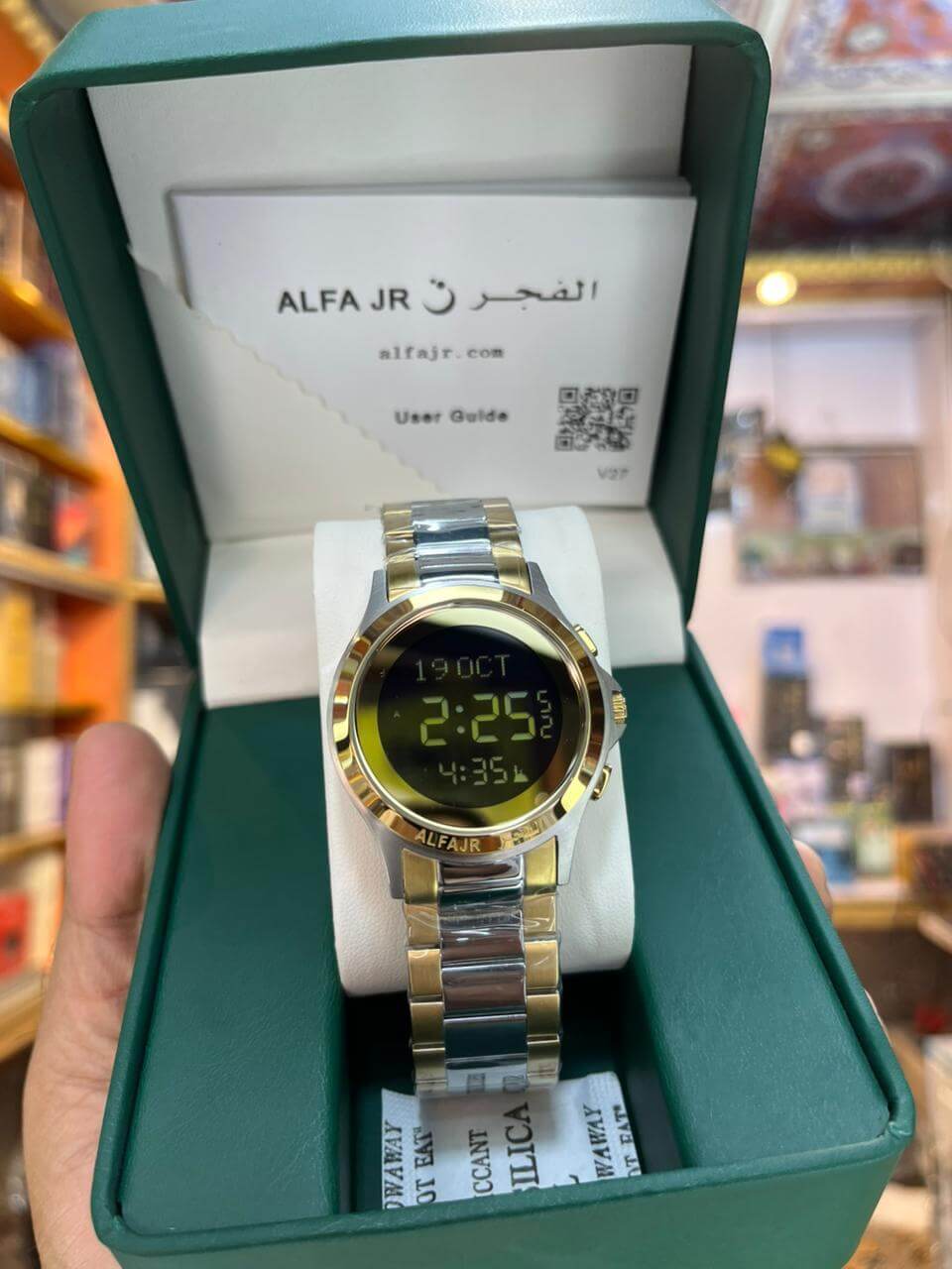 Digital watch with a gold and silver band in an open green box, displaying brand 'Alfa Jr'.