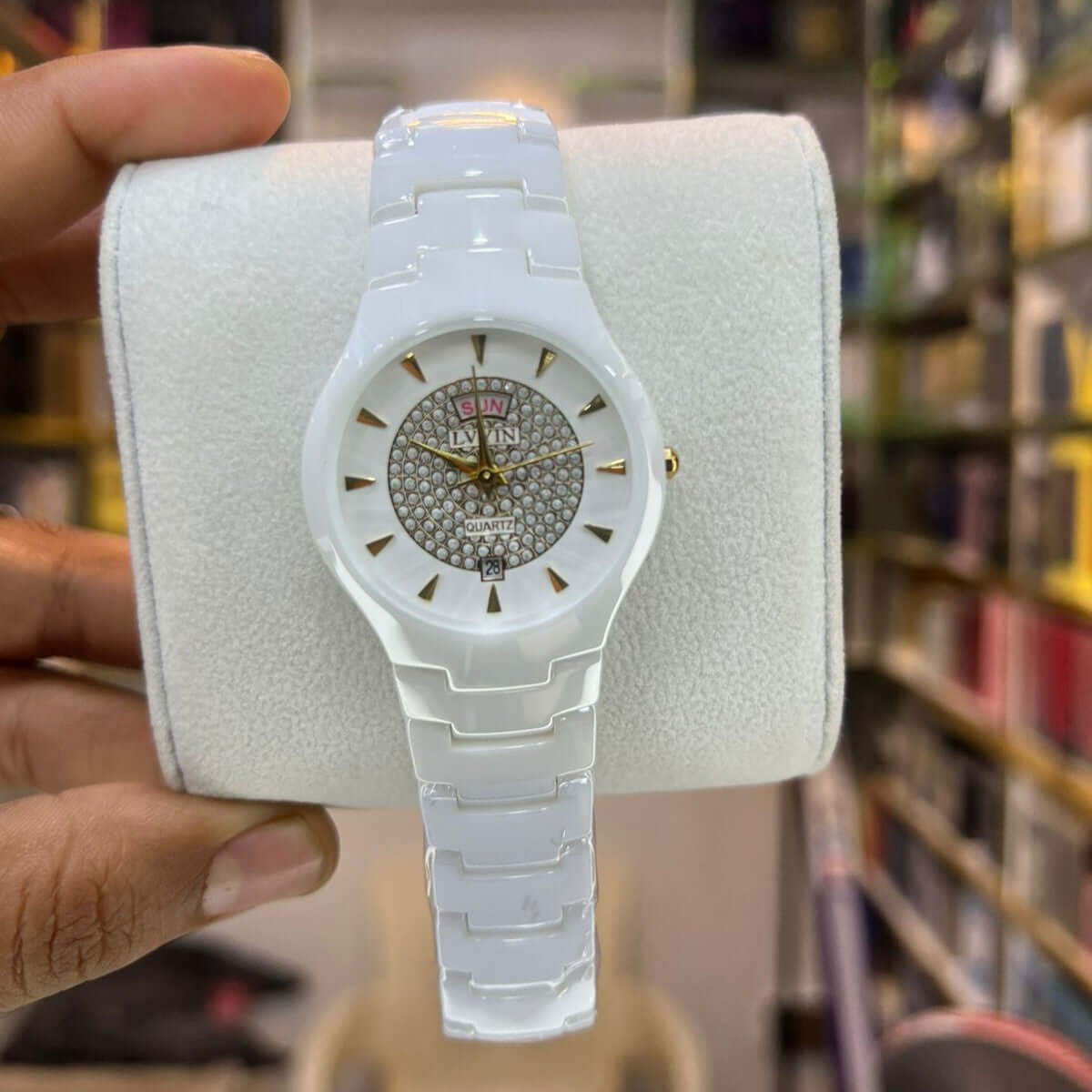 White watch with a decorative face held by a hand against a blurred bookshelf background